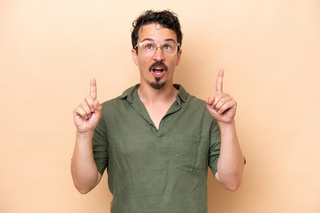 Young caucasian man isolated on beige background surprised and pointing up