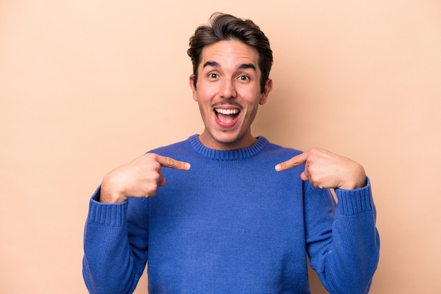 Young caucasian man isolated on beige background surprised pointing with finger, smiling broadly.