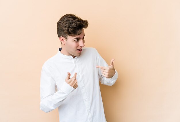 Young caucasian man isolated on beige background surprised pointing with finger, smiling broadly.