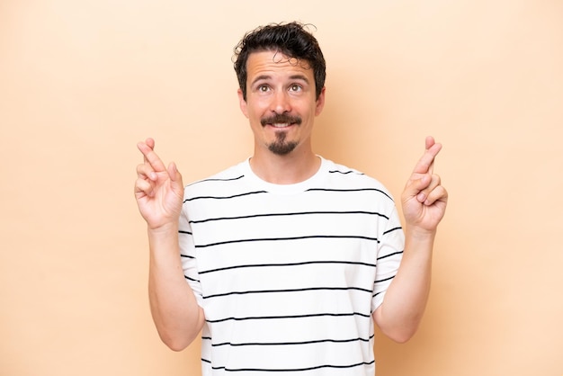 young caucasian man isolated on beige background with fingers crossing and wishing the best