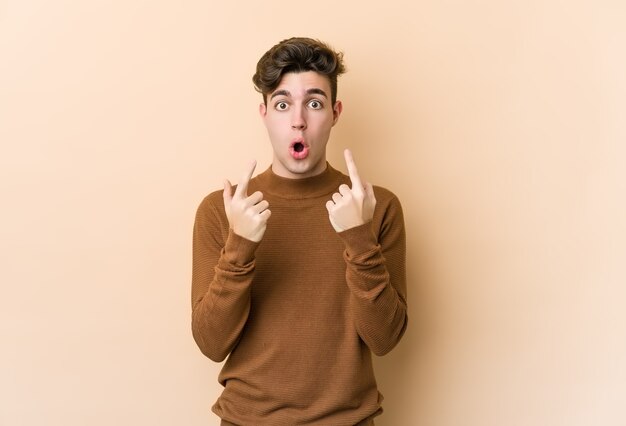 Young caucasian man isolated on beige wall pointing upside with opened mouth.