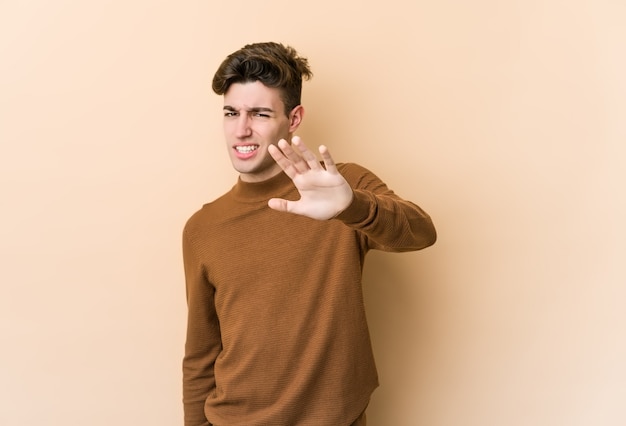 Young caucasian man isolated on beige wall rejecting someone showing a gesture of disgust.