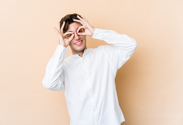 Young caucasian man isolated on beige wall showing okay sign over eyes