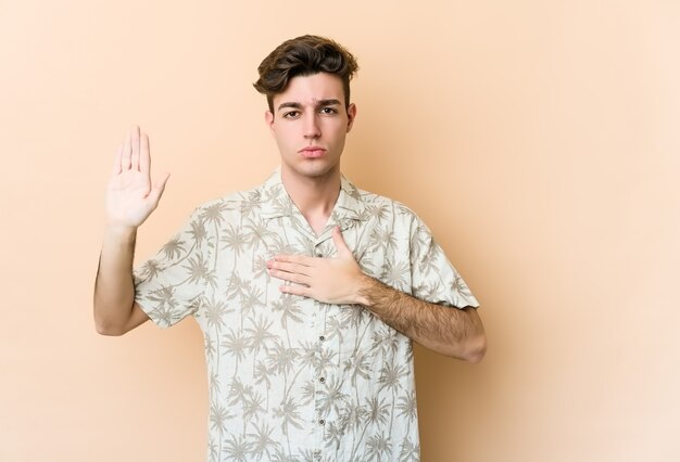 Young caucasian man isolated on beige wall taking an oath, putting hand on chest.
