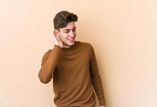 Young caucasian man isolated on beige wall trying to listening a gossip.