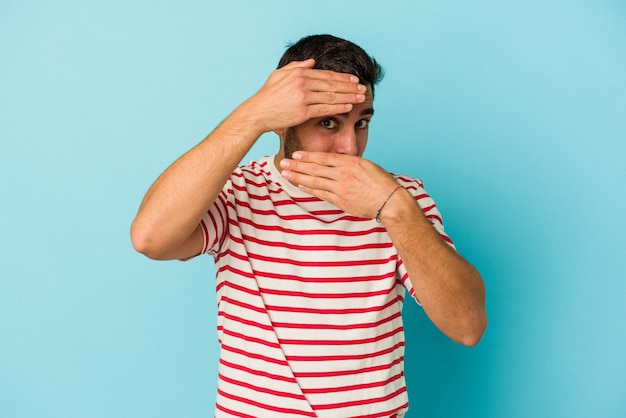 Young caucasian man isolated on blue background blink at the camera through fingers, embarrassed covering face.