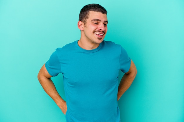 Young caucasian man isolated on blue background confident keeping hands on hips.