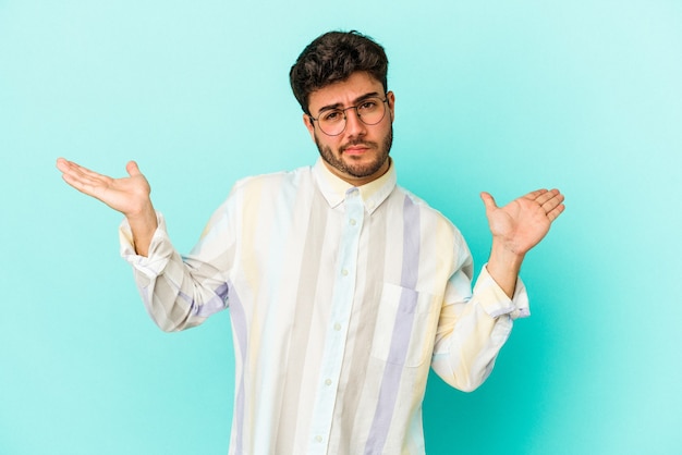 Young caucasian man isolated on blue background confused and doubtful shrugging shoulders to hold a copy space