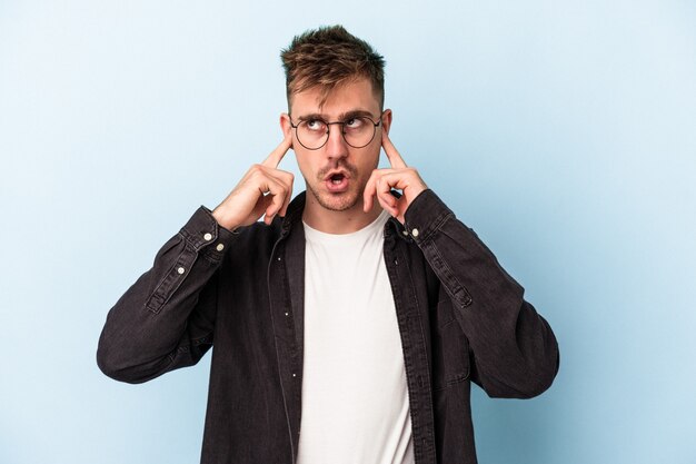 Young caucasian man isolated on blue background covering ears with fingers, stressed and desperate by a loudly ambient.