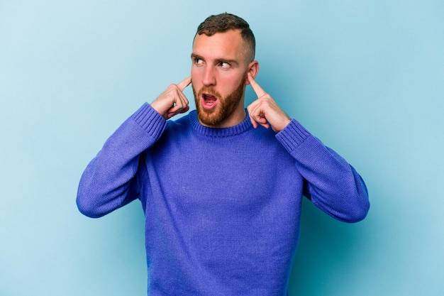 Young caucasian man isolated on blue background covering ears with fingers, stressed and desperate by a loudly ambient.