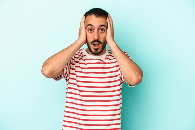 Young caucasian man isolated on blue background covering ears with hands trying not to hear too loud sound.