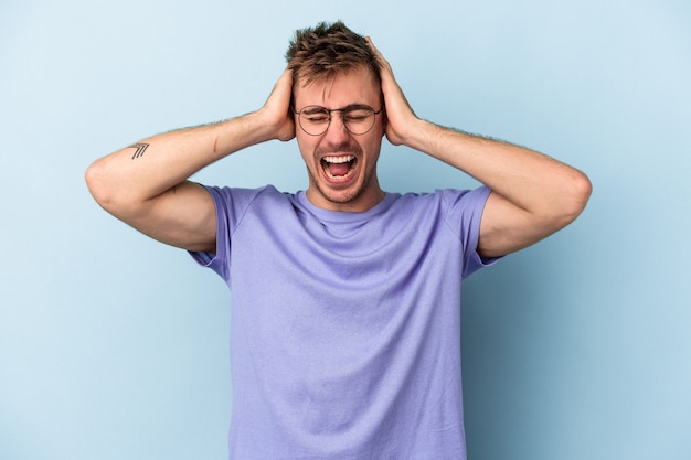 Young caucasian man isolated on blue background covering ears with hands trying not to hear too loud sound.