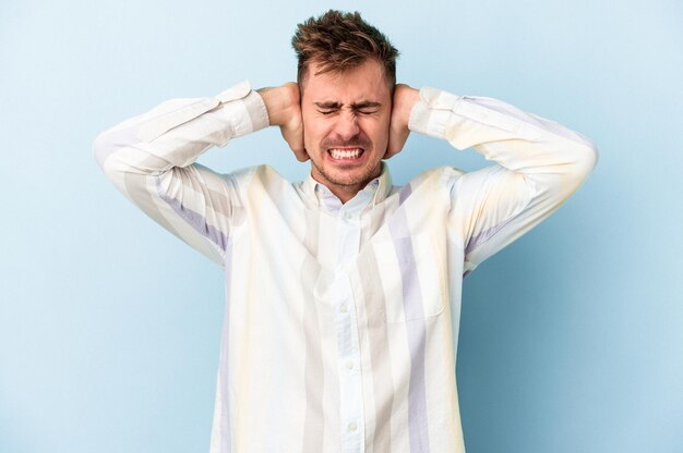 Young caucasian man isolated on blue background covering ears with hands.