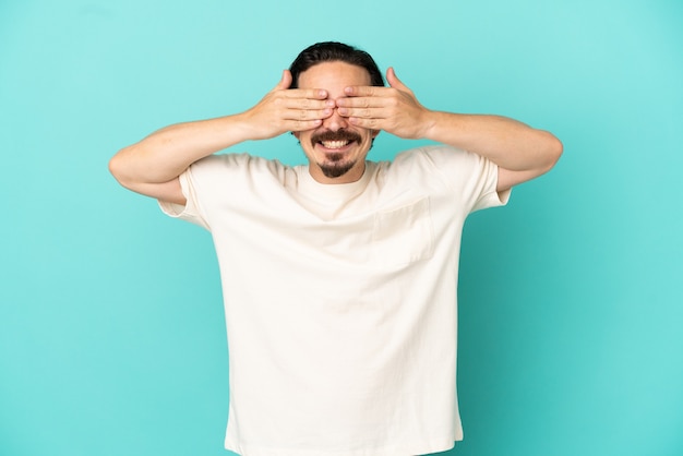 Young caucasian man isolated on blue background covering eyes by hands and smiling