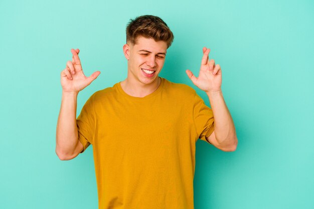 Young caucasian man isolated on blue background crossing fingers for having luck