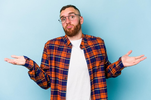 Young caucasian man isolated on blue background doubting and shrugging shoulders in questioning gesture.