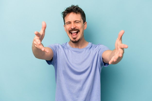 Young caucasian man isolated on blue background feels confident giving a hug to the camera.