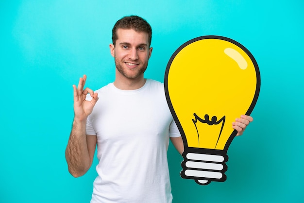 Young caucasian man isolated on blue background holding a bulb icon and doing OK sign