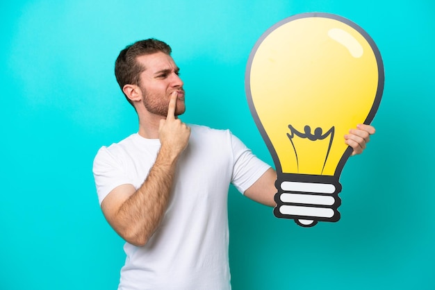 Young caucasian man isolated on blue background holding a bulb icon and having doubts