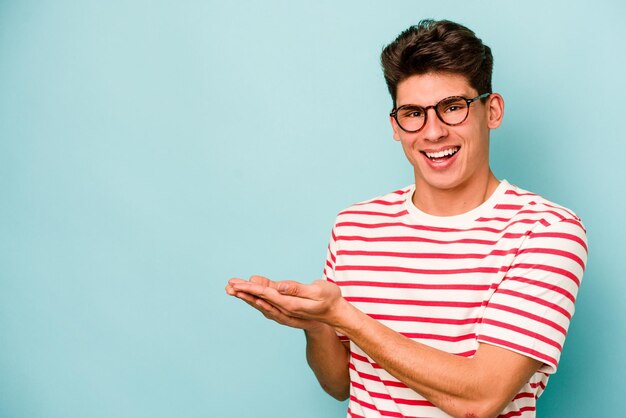 Young caucasian man isolated on blue background holding a copy space on a palm