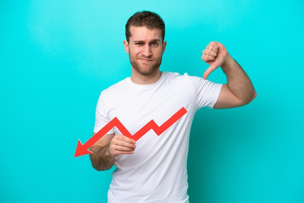 Young caucasian man isolated on blue background holding a\
downward arrow and doing bad signal