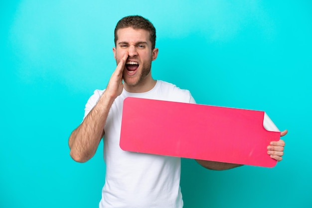 Young caucasian man isolated on blue background holding an empty placard and shouting