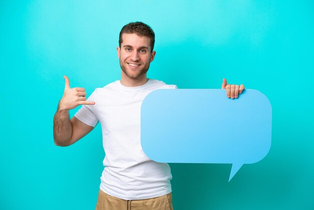 Young caucasian man isolated on blue background holding an empty speech bubble and doing phone gesture