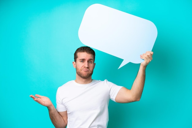 Young caucasian man isolated on blue background holding an empty speech bubble and having doubts