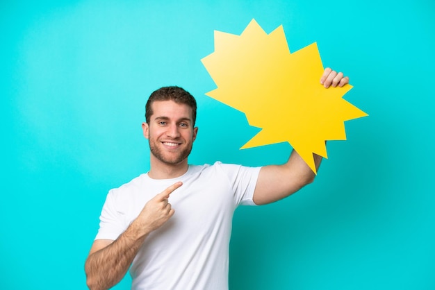 Young caucasian man isolated on blue background holding an empty speech bubble and pointing it