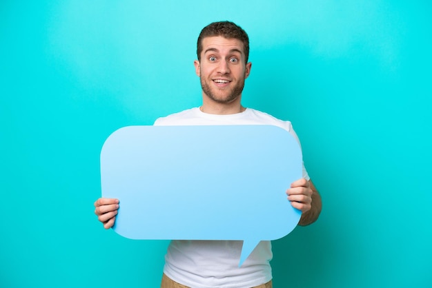 Young caucasian man isolated on blue background holding an empty speech bubble with surprised expression