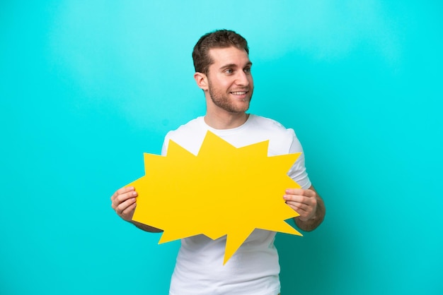 Young caucasian man isolated on blue background holding an empty speech bubble