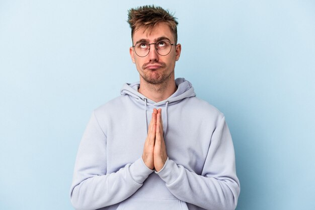 Young caucasian man isolated on blue background holding hands in pray near mouth, feels confident.