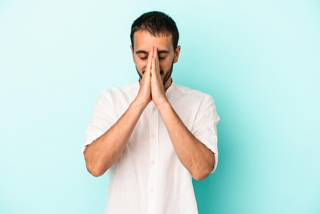 Young caucasian man isolated on blue background holding hands in pray near mouth feels confident