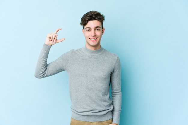 Young caucasian man isolated on blue background holding something little with forefingers, smiling and confident.