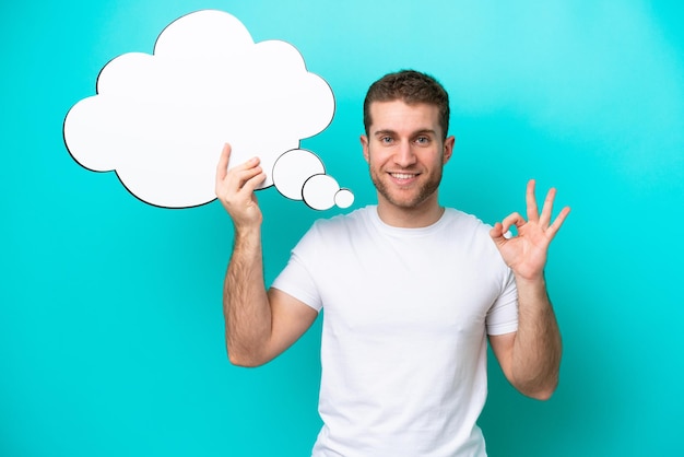 Young caucasian man isolated on blue background holding a thinking speech bubble and doing OK sign