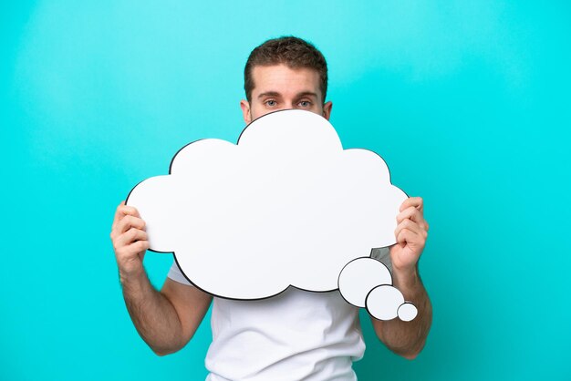 Young caucasian man isolated on blue background holding a thinking speech bubble and hiding behind it