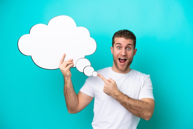 Young caucasian man isolated on blue background holding a thinking speech bubble with surprised expression