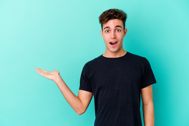 Young caucasian man isolated on blue background impressed holding copy space on palm.