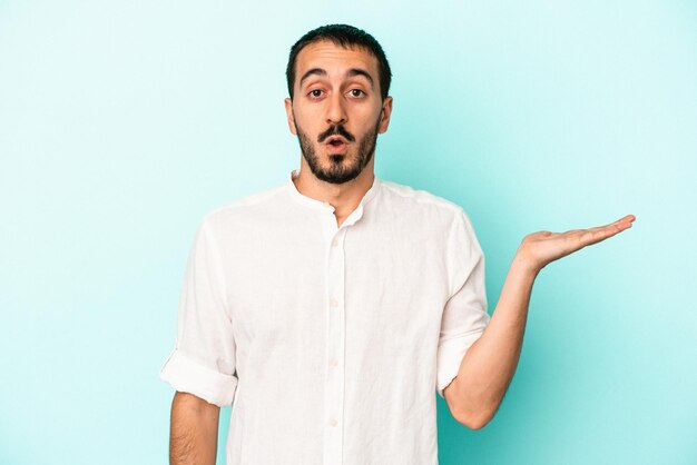 Young caucasian man isolated on blue background impressed holding copy space on palm.