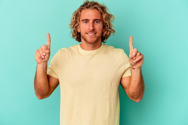 Young caucasian man isolated on blue background indicates with both fore fingers up showing a blank space.