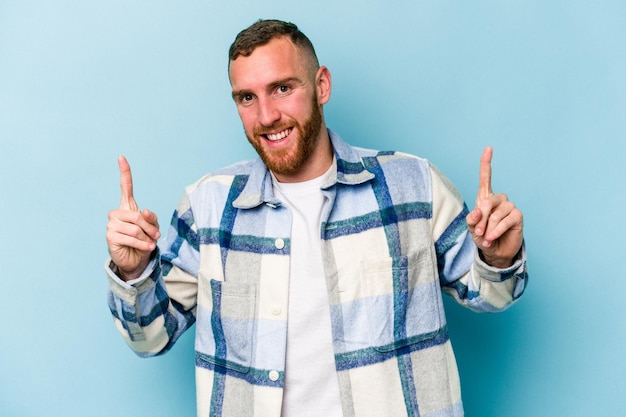 Young caucasian man isolated on blue background indicates with both fore fingers up showing a blank space.