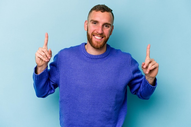 Young caucasian man isolated on blue background indicates with both fore fingers up showing a blank space