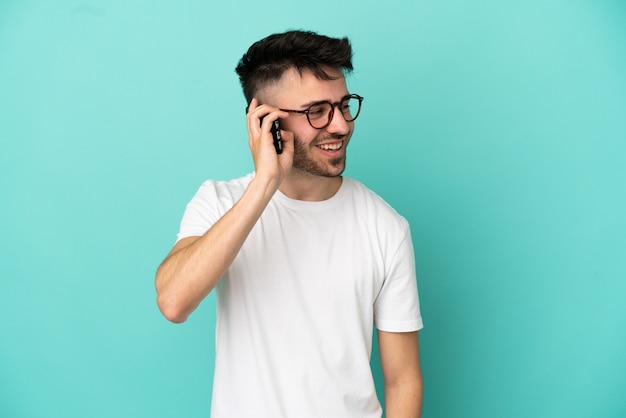 Young caucasian man isolated on blue background keeping a conversation with the mobile phone with someone