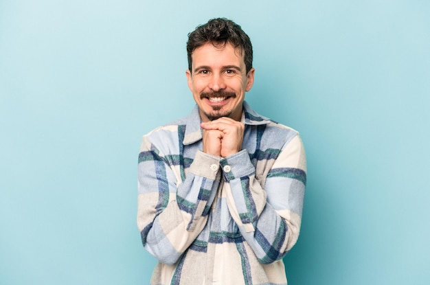 Young caucasian man isolated on blue background keeps hands under chin, is looking happily aside.