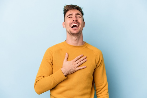 Il giovane uomo caucasico isolato su sfondo blu ride ad alta voce tenendo la mano sul petto.