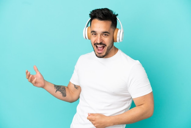 Giovane uomo caucasico isolato su sfondo blu ascoltando musica e facendo il gesto della chitarra