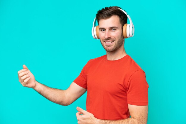 Photo young caucasian man isolated on blue background listening music and doing guitar gesture