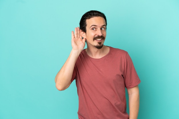 Foto giovane uomo caucasico isolato su sfondo blu ascoltando qualcosa mettendo la mano sull'orecchio