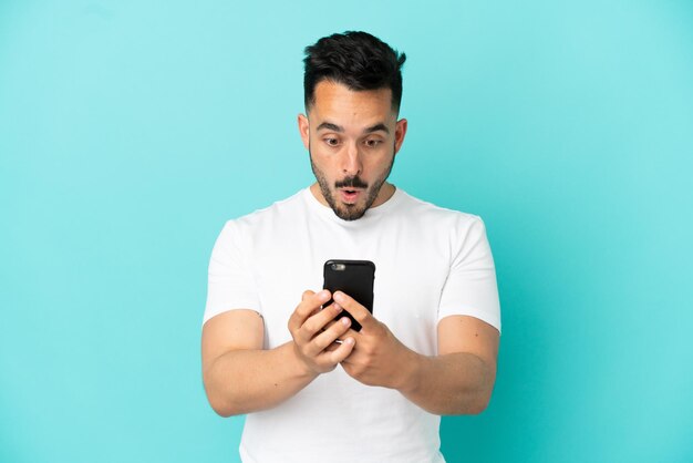 Young caucasian man isolated on blue background looking at the camera while using the mobile with surprised expression