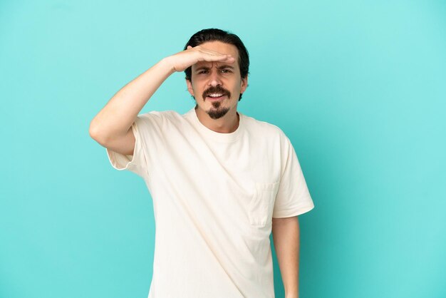 Young caucasian man isolated on blue background looking far away with hand to look something